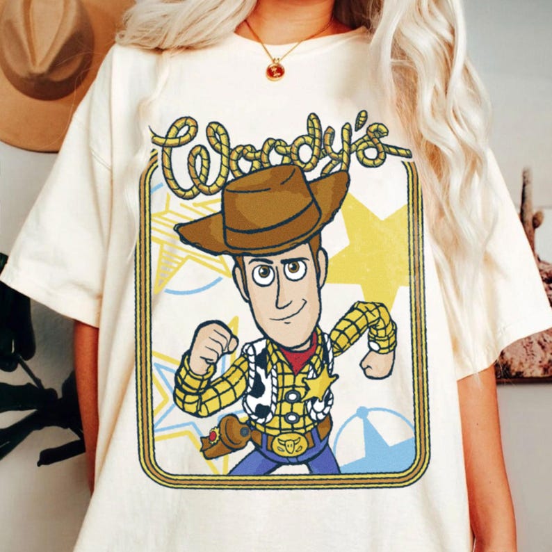 May include: A cream-colored short-sleeve t-shirt featuring a graphic print of a cowboy character wearing a brown hat, yellow plaid shirt, and cow-print vest. The text Woody's is written in a rope-style font above the character inside a border.