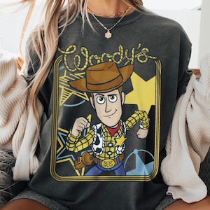 May include: A charcoal gray crewneck sweatshirt featuring a graphic print of the character Woody from Toy Story. The design includes the text Woody's in a rope-style font, a yellow star background, and a framed border with a western aesthetic.
