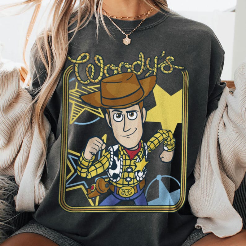May include: A charcoal gray crewneck sweatshirt featuring a graphic print of the character Woody from Toy Story. The design includes the text Woody's in a rope-style font, a yellow star background, and a framed border with a western aesthetic.