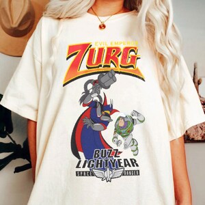 May include: Cream-colored t-shirt featuring a graphic of Zurg, Buzz Lightyear, and the text "EVIL EMPEROR ZURG" and "BUZZ LIGHTYEAR SPACE RANGER". The design includes red, orange, blue, and green colors.