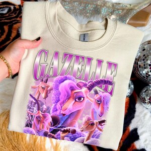 May include: Cream-colored sweatshirt featuring a graphic of cartoon gazelles with purple hair and accents. The word "GAZELLE" is printed in a purple, outlined font above the gazelles. The sweatshirt is held by a person.