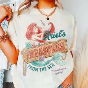 May include: Cream-colored t-shirt featuring a graphic of Ariel from The Little Mermaid. The design includes Ariel, a wooden sign that reads "Ariel's Treasures From The Sea", and the phrase "Dinglehoppers aplenty!"