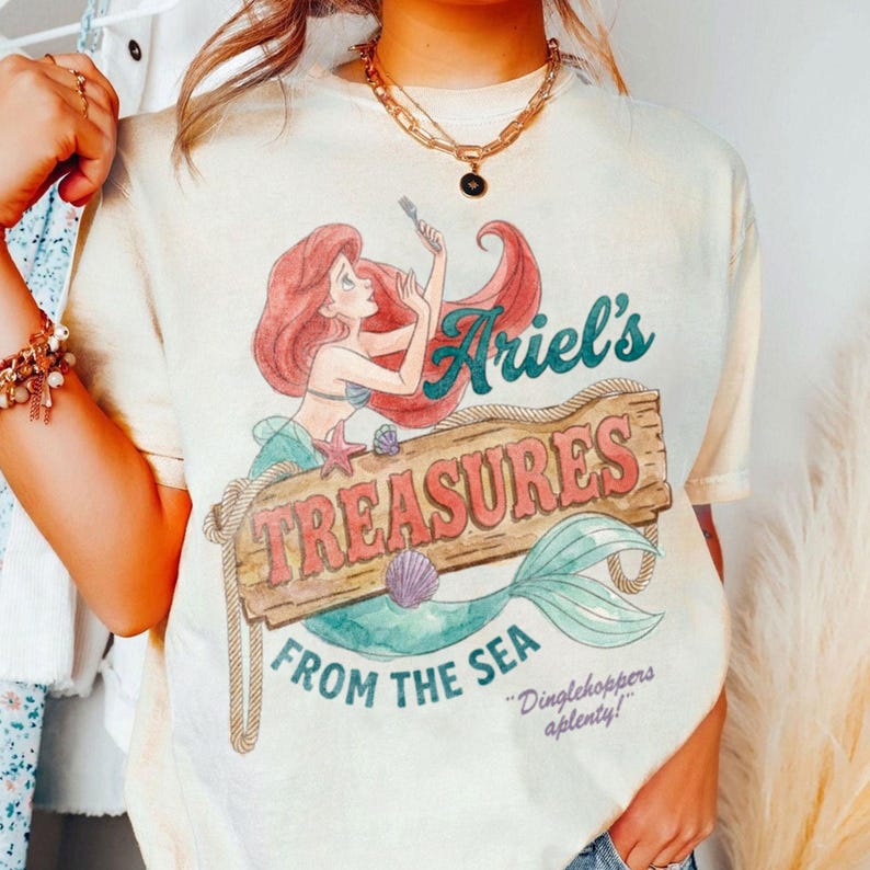May include: Cream-colored t-shirt featuring a graphic of Ariel from The Little Mermaid. The design includes Ariel, a wooden sign that reads "Ariel's Treasures From The Sea", and the phrase "Dinglehoppers aplenty!"