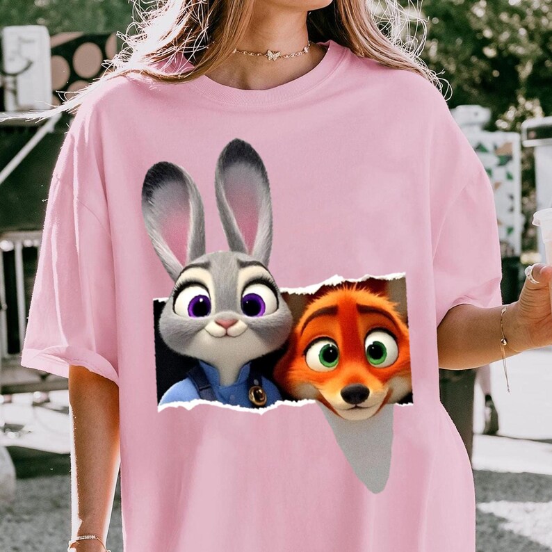 May include: A light pink t-shirt featuring a graphic of Judy Hopps and Nick Wilde from the animated movie Zootopia. Judy, a gray rabbit, and Nick, an orange fox, are depicted peeking through a torn paper effect.
