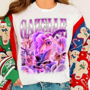May include: White t-shirt featuring a purple graphic of a gazelle with purple hair and the word "GAZELLE" in a purple, outlined font. The gazelle is holding a microphone. The shirt is worn with a Christmas sweater.