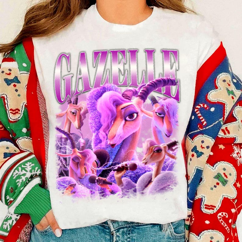 May include: White t-shirt featuring a purple graphic of a gazelle with purple hair and the word "GAZELLE" in a purple, outlined font. The gazelle is holding a microphone. The shirt is worn with a Christmas sweater.
