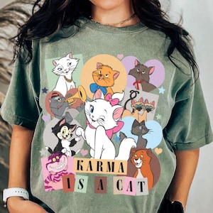 Puede incluir: Camiseta verde con una ilustraci&oacute;n de dibujos animados de varios gatos. El texto "KARMA IS A CAT" est&aacute; impreso en la camiseta.