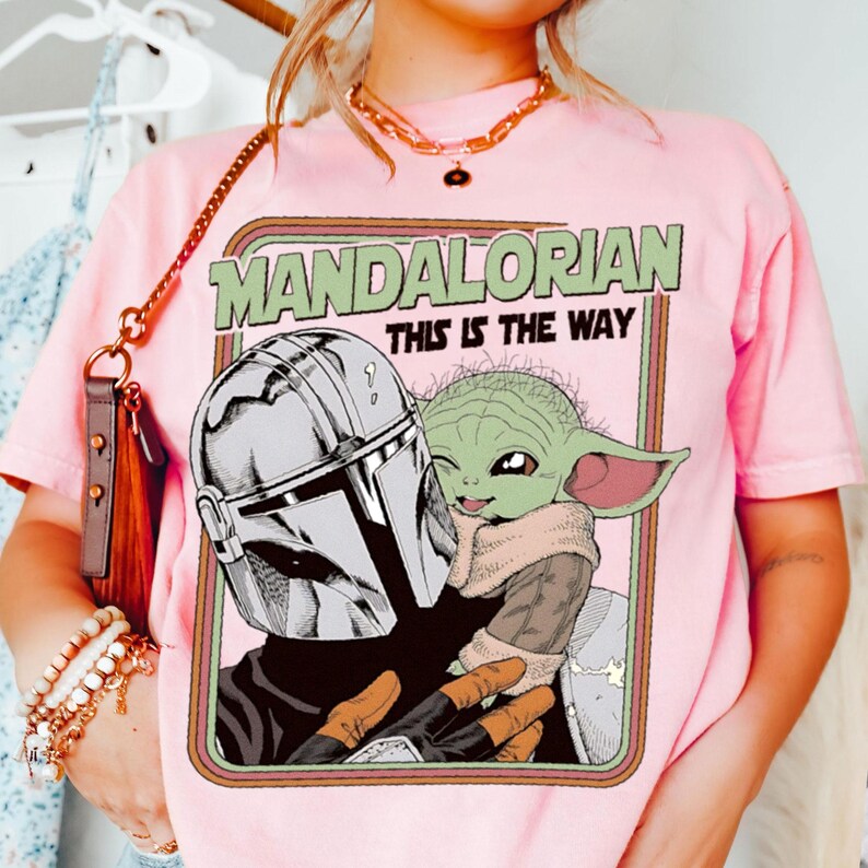 Puede incluir: Camiseta rosa con un gr&aacute;fico de The Mandalorian y Grogu, con el texto "MANDALORIAN THIS IS THE WAY" en un marco retro. La ilustraci&oacute;n muestra el casco del Mandaloriano y la cara de Grogu. La camiseta tiene mangas cortas.