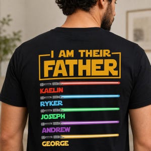 May include: Black t-shirt featuring the text "I AM THEIR FATHER" in yellow. Below the text are six coloured lightsabers with names: Kaelin, Ryker, Joseph, Andrew, and George. The lightsabers are red, blue, green, purple, and yellow.