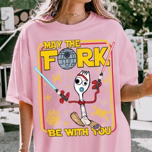 May include: Pink t-shirt with a graphic featuring a fork character holding lightsabers, a Death Star, and the text "MAY THE FORK BE WITH YOU." The design is framed in yellow and red.