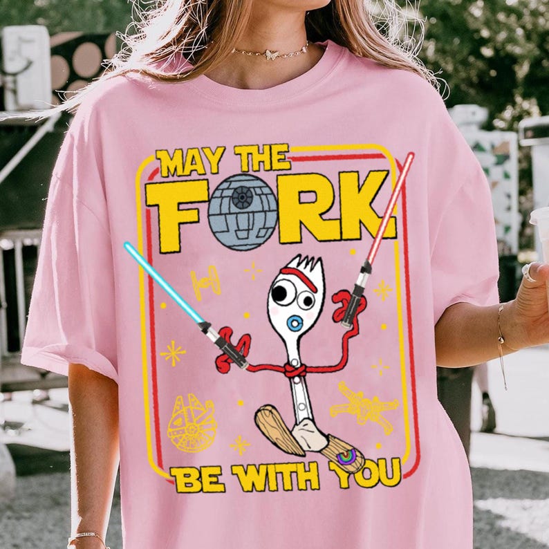May include: Pink t-shirt with a graphic featuring a fork character holding lightsabers, a Death Star, and the text "MAY THE FORK BE WITH YOU." The design is framed in yellow and red.