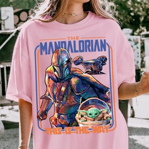 May include: Pink t-shirt featuring a colorful graphic of The Mandalorian and Grogu from the Star Wars series. The design includes the text "THE MANDALORIAN" and "THIS IS THE WAY" in a blue and orange frame.