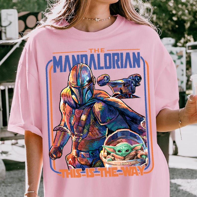 May include: Pink t-shirt featuring a colorful graphic of The Mandalorian and Grogu from the Star Wars series. The design includes the text "THE MANDALORIAN" and "THIS IS THE WAY" in a blue and orange frame.