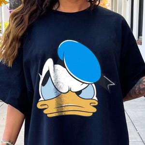 May include: Navy blue t-shirt featuring a distressed graphic of Donald Duck's face. The cartoon character's face is white, yellow, and light blue, with a blue sailor hat. The shirt is a casual style.