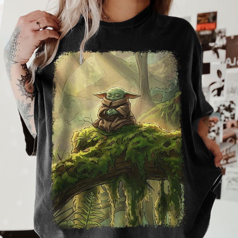 May include: Black t-shirt with a detailed illustration of a green alien-like character in a brown robe, meditating on a moss-covered log in a forest. The artwork has a soft, painterly style, with a focus on nature.