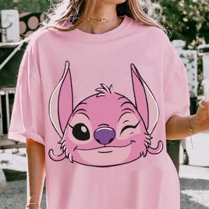 May include: Pink t-shirt featuring a graphic of a winking, pink cartoon character with large ears and a purple nose. The character's face is outlined in black. The shirt has short sleeves and a relaxed fit.