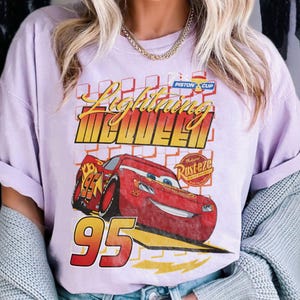 May include: A light purple t-shirt featuring Lightning McQueen from the movie Cars. The graphic includes the text "Lightning McQueen" in yellow and red, a red race car, and the number "95".