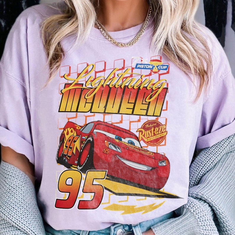 May include: A light purple t-shirt featuring Lightning McQueen from the movie Cars. The graphic includes the text "Lightning McQueen" in yellow and red, a red race car, and the number "95".