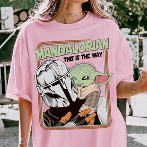 Puede incluir: Camiseta rosa claro con un dise&ntilde;o de The Mandalorian y Grogu de la serie Star Wars. El dise&ntilde;o incluye el texto "MANDALORIAN THIS IS THE WAY" en verde y naranja. La obra de arte representa a los personajes en un estilo retro.