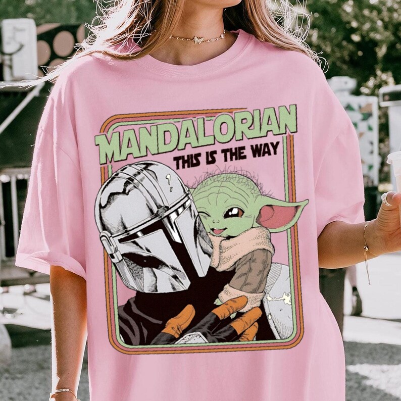 Puede incluir: Camiseta rosa claro con un dise&ntilde;o de The Mandalorian y Grogu de la serie Star Wars. El dise&ntilde;o incluye el texto "MANDALORIAN THIS IS THE WAY" en verde y naranja. La obra de arte representa a los personajes en un estilo retro.