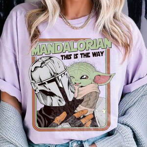 Puede incluir: Camiseta lila claro con un gr&aacute;fico de The Mandalorian y Grogu. El dise&ntilde;o incluye el texto "MANDALORIAN THIS IS THE WAY" en verde y naranja. La obra de arte representa el casco mandaloriano y Grogu en un estilo retro.