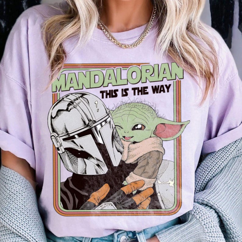 Puede incluir: Camiseta lila claro con un gr&aacute;fico de The Mandalorian y Grogu. El dise&ntilde;o incluye el texto "MANDALORIAN THIS IS THE WAY" en verde y naranja. La obra de arte representa el casco mandaloriano y Grogu en un estilo retro.
