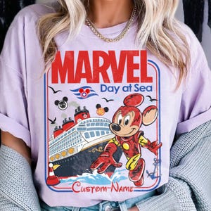 May include: A light purple t-shirt featuring a Marvel Day at Sea design. The graphic includes a cruise ship, Mickey Mouse in an Iron Man suit, and the text "MARVEL Day at Sea". The design has a vintage, cartoon style.