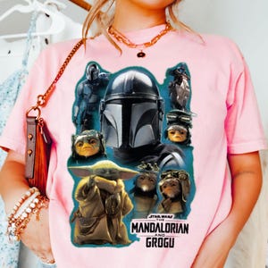 May include: Pale pink t-shirt with a graphic featuring characters from The Mandalorian series, including the Mandalorian, Grogu, and other creatures. The Star Wars logo is also visible. The shirt is a light pink colour.