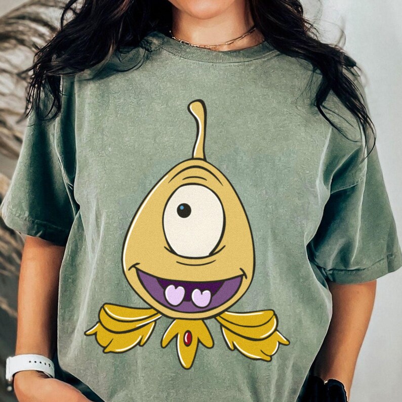 May include: Sage green t-shirt featuring a cartoon character with a single eye, yellow body, and purple heart-shaped teeth. The character has a smiling expression and is centered on the shirt.