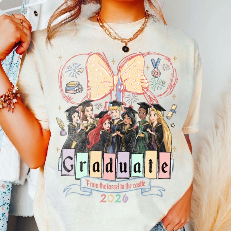 Puede incluir: Camiseta color crema con un gr&aacute;fico de princesas de Disney con atuendo de graduaci&oacute;n. El dise&ntilde;o incluye un gran lazo, s&iacute;mbolos de graduaci&oacute;n y el texto "Graduate From the tassel to the castle 2026".