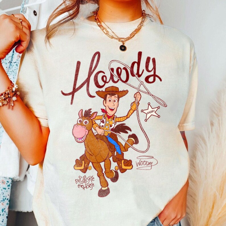 May include: Cream-colored t-shirt featuring a cartoon image of Woody from Toy Story riding Bullseye, with the word "Howdy" in a brown script above. The shirt also includes the words "Woody" and "Bullseye the Horse".