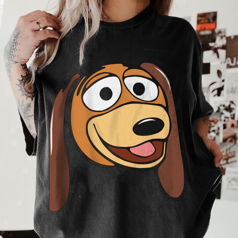 Pu&ograve; includere: T-shirt nero con un'immagine a cartoni animati di Slinky Dog di Toy Story. La grafica mostra il volto di Slinky con grandi occhi bianchi, un muso marrone chiaro e orecchie marroni. La maglietta &egrave; di colore scuro e sembra essere fatta di materiale morbido.