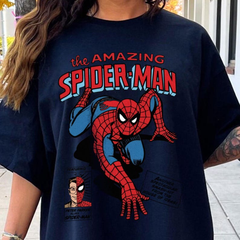 以下が含まれることがあります： ネイビーブルーのTシャツ。コミック風のスパイダーマンのイラストが特徴です。「the AMAZING SPIDER-MAN」の文字が赤と青で描かれ、ピーター・パーカーの画像とテキストも含まれています。