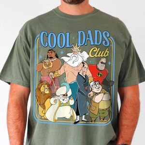 May include: Olive green t-shirt with a cartoon graphic of animated characters and the text "COOL DADS Club" in blue. The graphic includes a lion, a character with a trident, and other figures.