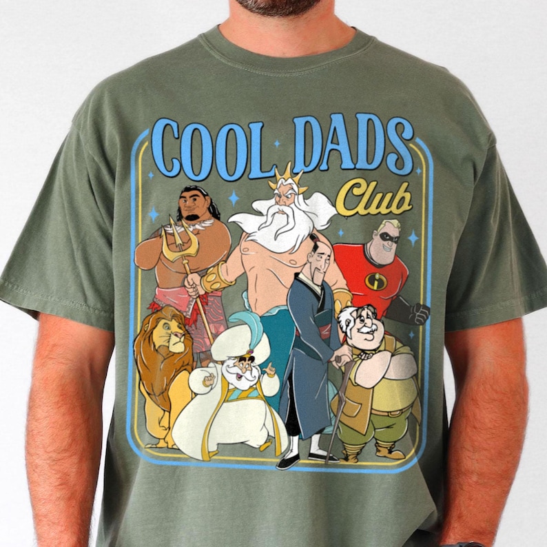 May include: Olive green t-shirt with a cartoon graphic of animated characters and the text "COOL DADS Club" in blue. The graphic includes a lion, a character with a trident, and other figures.