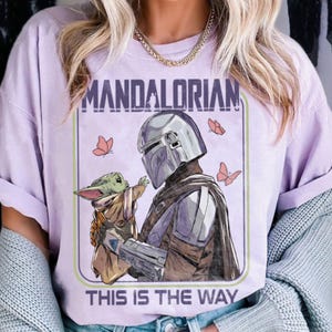 May include: A light purple t-shirt featuring a graphic of The Mandalorian and Grogu, with the text "MANDALORIAN" above and "THIS IS THE WAY" below. The design includes butterflies and a green and purple border.