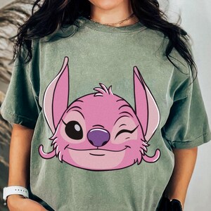 May include: Sage green t-shirt featuring a pink cartoon character with large ears, winking eye, and a purple nose. The character's face is centered on the shirt. The shirt has short sleeves and a relaxed fit.