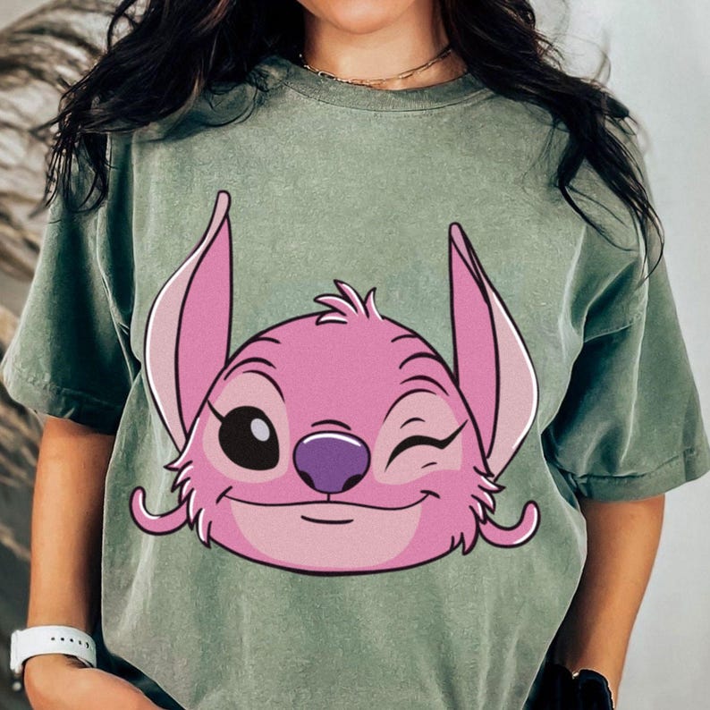 May include: Sage green t-shirt featuring a pink cartoon character with large ears, winking eye, and a purple nose. The character's face is centered on the shirt. The shirt has short sleeves and a relaxed fit.