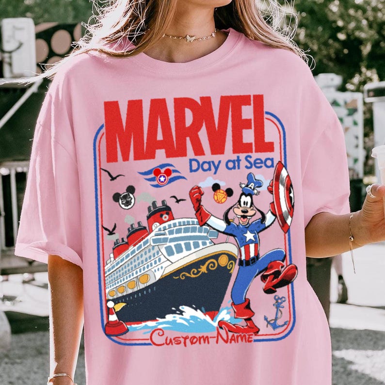 May include: Pink t-shirt featuring a Marvel Day at Sea design. The graphic includes the word "MARVEL" in red, a cruise ship, and a cartoon character dressed as Captain America. The shirt has a custom name area at the bottom.