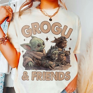 K&ouml;nnte beinhalten: Cremefarbenes T-Shirt mit dem Text "GROGU & FRIENDS" in braunen Buchstaben. Die Grafik zeigt Grogu und drei weitere au&szlig;erirdische Charaktere. Das Design hat ein Vintage-Aussehen, leicht verblasst.
