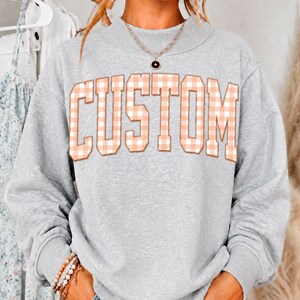 May include: A heather gray long-sleeve sweatshirt featuring the word CUSTOM in large, block-style letters with a light peach and white gingham check pattern. The garment has a crew neckline and is paired with a delicate gold-tone chain necklace.