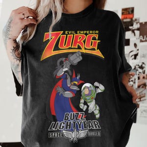 May include: Black t-shirt featuring a graphic of Evil Emperor Zurg and Buzz Lightyear from Toy Story. The design includes the text "EVIL EMPEROR ZURG" in yellow and red, and "BUZZ LIGHTYEAR SPACE RANGER" below the characters.