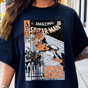 May include: Navy blue t-shirt featuring a comic book cover of Spider-Man. The graphic includes the text "the AMAZING SPIDER-MAN" and "ESCAPE IMPOSSIBLE!". The comic book cover is in orange, black, white, and gray.