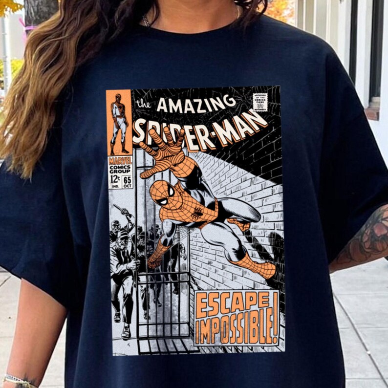 May include: Navy blue t-shirt featuring a comic book cover of Spider-Man. The graphic includes the text "the AMAZING SPIDER-MAN" and "ESCAPE IMPOSSIBLE!". The comic book cover is in orange, black, white, and gray.