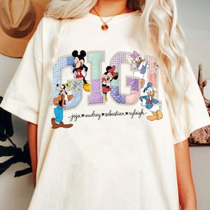 May include: Cream-coloured t-shirt with the word "GIGI" in a patchwork design, featuring Disney characters. Mickey Mouse, Minnie Mouse, Goofy, Donald Duck, and Daisy Duck are included. The names "jojo audrey sebastian ryleigh" are printed below.