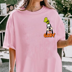 Puede incluir: Una camiseta rosa claro con una imagen de dibujos animados de Goofy. La imagen muestra la cara y el sombrero de Goofy, con la palabra "GOOFY" en letras naranjas debajo. La camiseta tiene mangas cortas y un corte holgado.