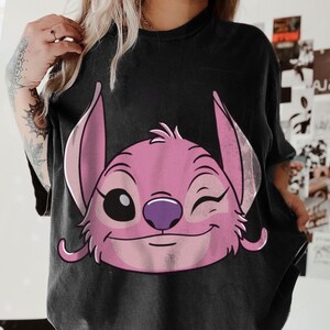 May include: Black t-shirt featuring a pink cartoon character with large ears, a purple nose, and a winking eye. The character's face is centered on the shirt. The shirt has short sleeves and a crew neck.