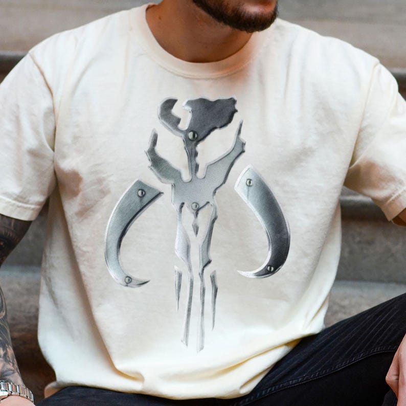 Puede incluir: Camiseta color crema con un emblema plateado met&aacute;lico. El emblema es un dise&ntilde;o sim&eacute;trico estilizado con elementos curvos y angulares, que recuerda a un casco o m&aacute;scara. La camiseta es una prenda casual.