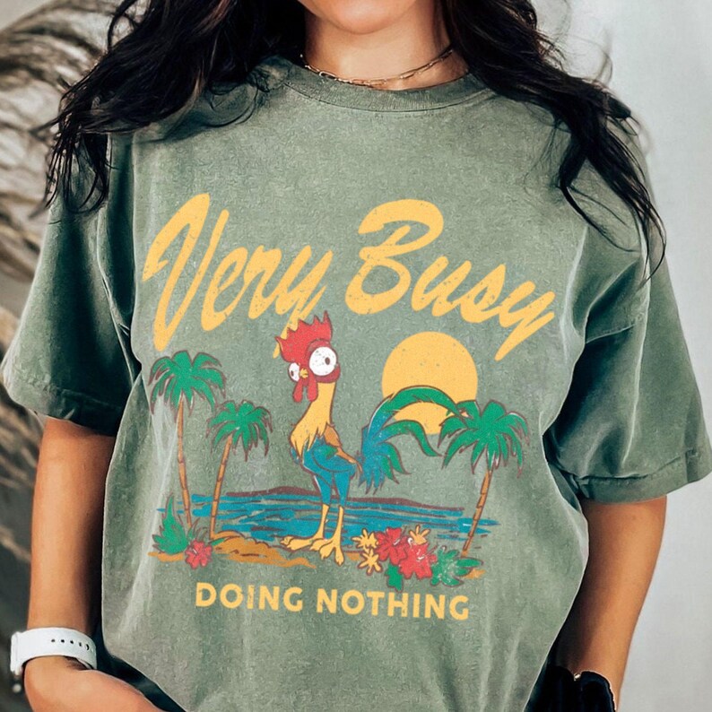 K&ouml;nnte beinhalten: Olivgr&uuml;nes T-Shirt mit dem Text "Very Busy Doing Nothing" in Gelb. Die Grafik zeigt einen Cartoon-Hahn, Palmen, einen Sonnenuntergang und Meereswellen. Das Shirt hat kurze &Auml;rmel.
