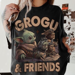 K&ouml;nnte beinhalten: Schwarzes T-Shirt mit einer Grafik von Grogu und Freunden. Das Design zeigt Grogu und drei weitere Charaktere mit dem Text "GROGU & FRIENDS" in einer verblichenen, braunen Schriftart. Das T-Shirt hat einen Vintage-Look.
